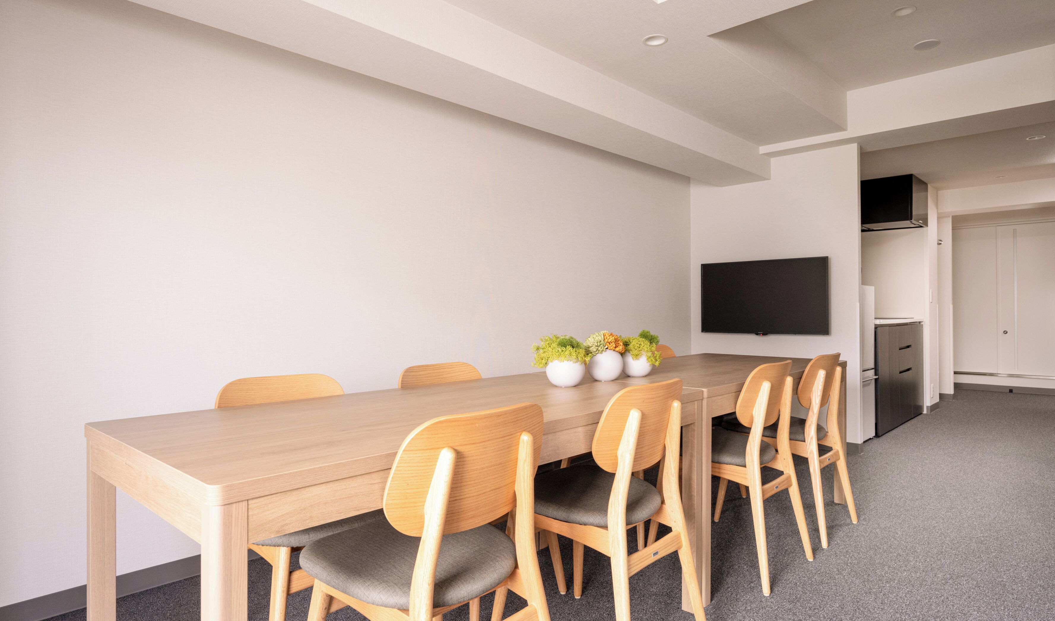 A modern meeting room with a long wooden table, six wooden chairs, and a flat-screen TV mounted on the wall.