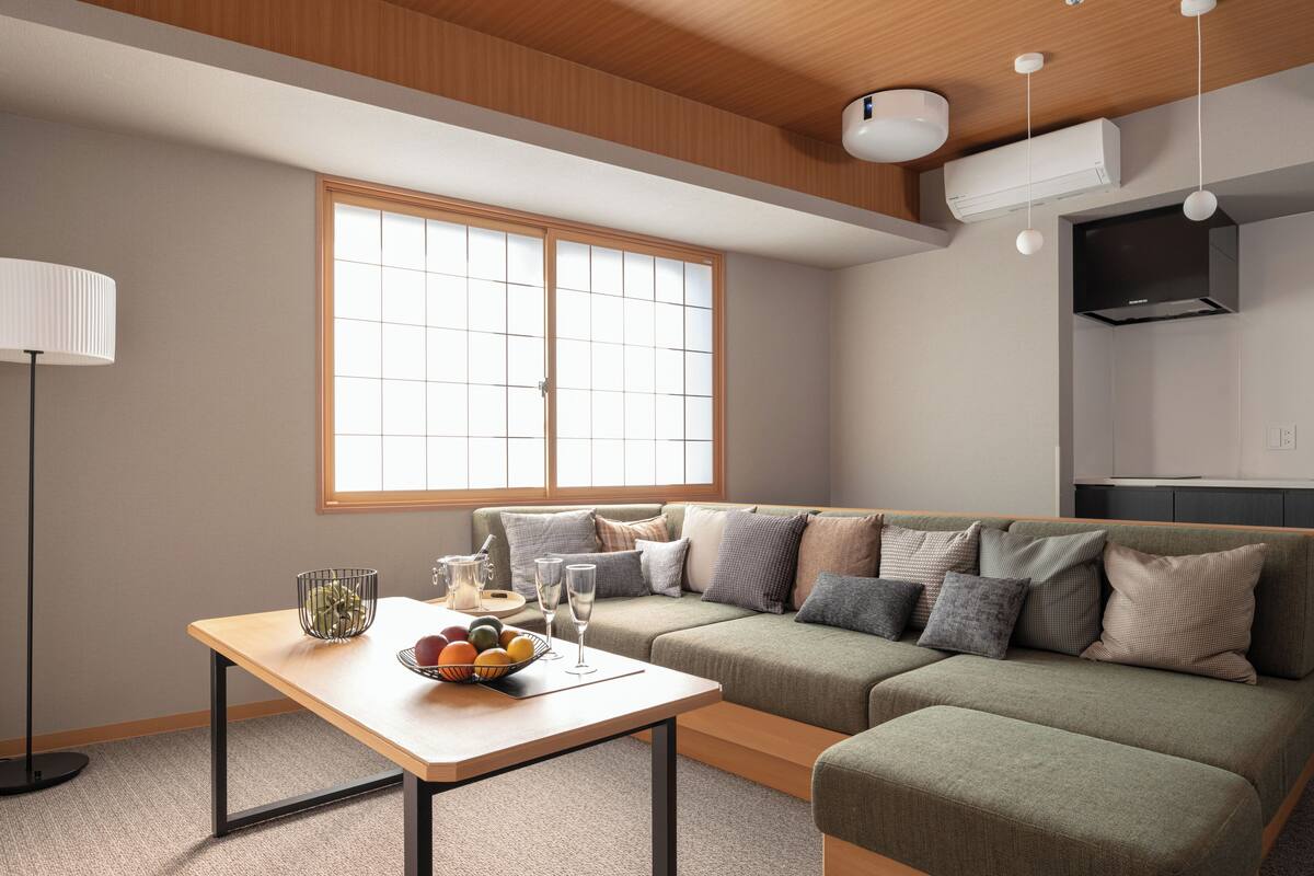 A modern living room with a sofa, a table with fruit and glasses, a lamp, and a large window.