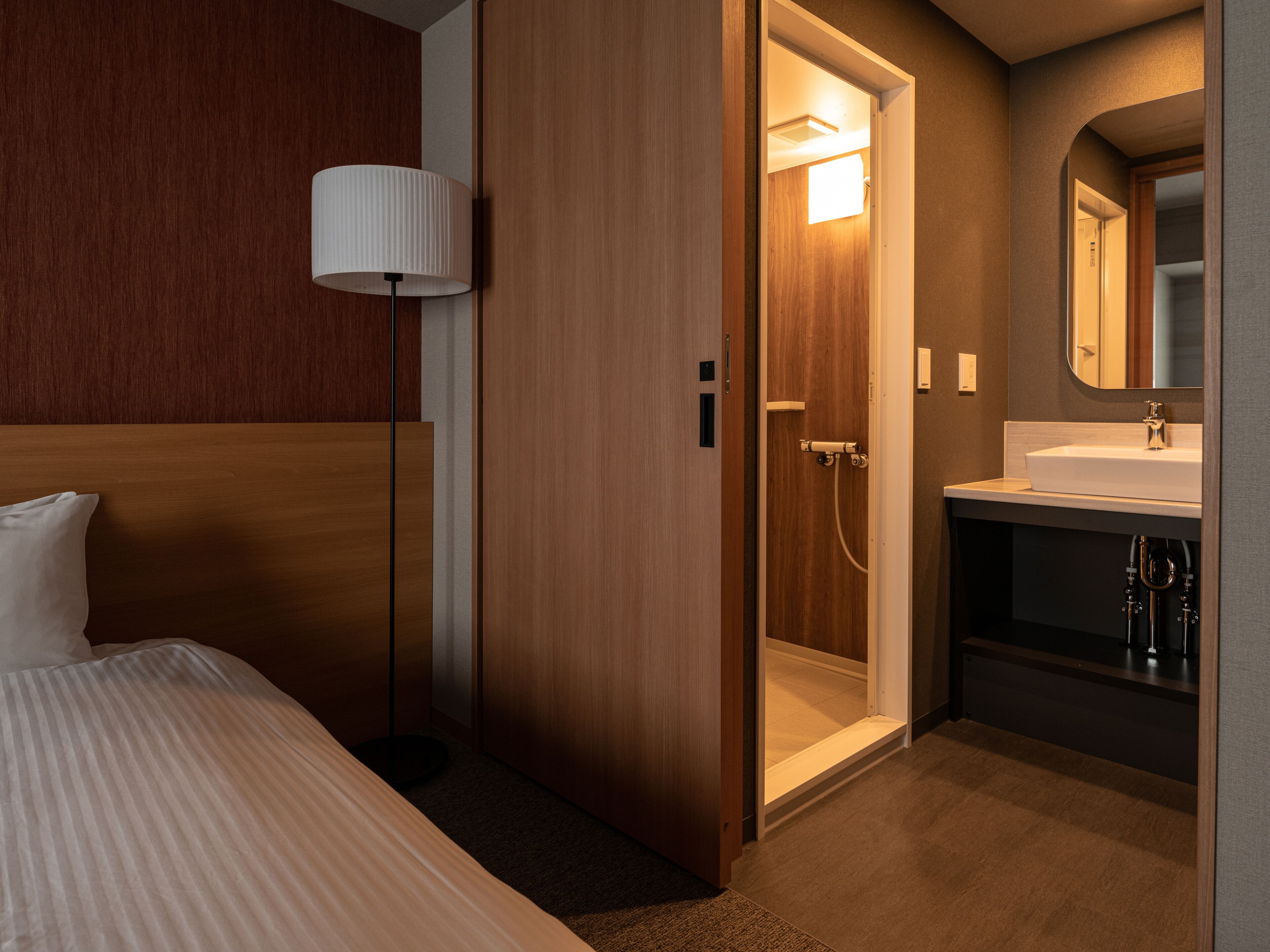 A hotel room with a bed, a bedside lamp, a wooden wardrobe, a shower area, and a bathroom with a sink and mirror.