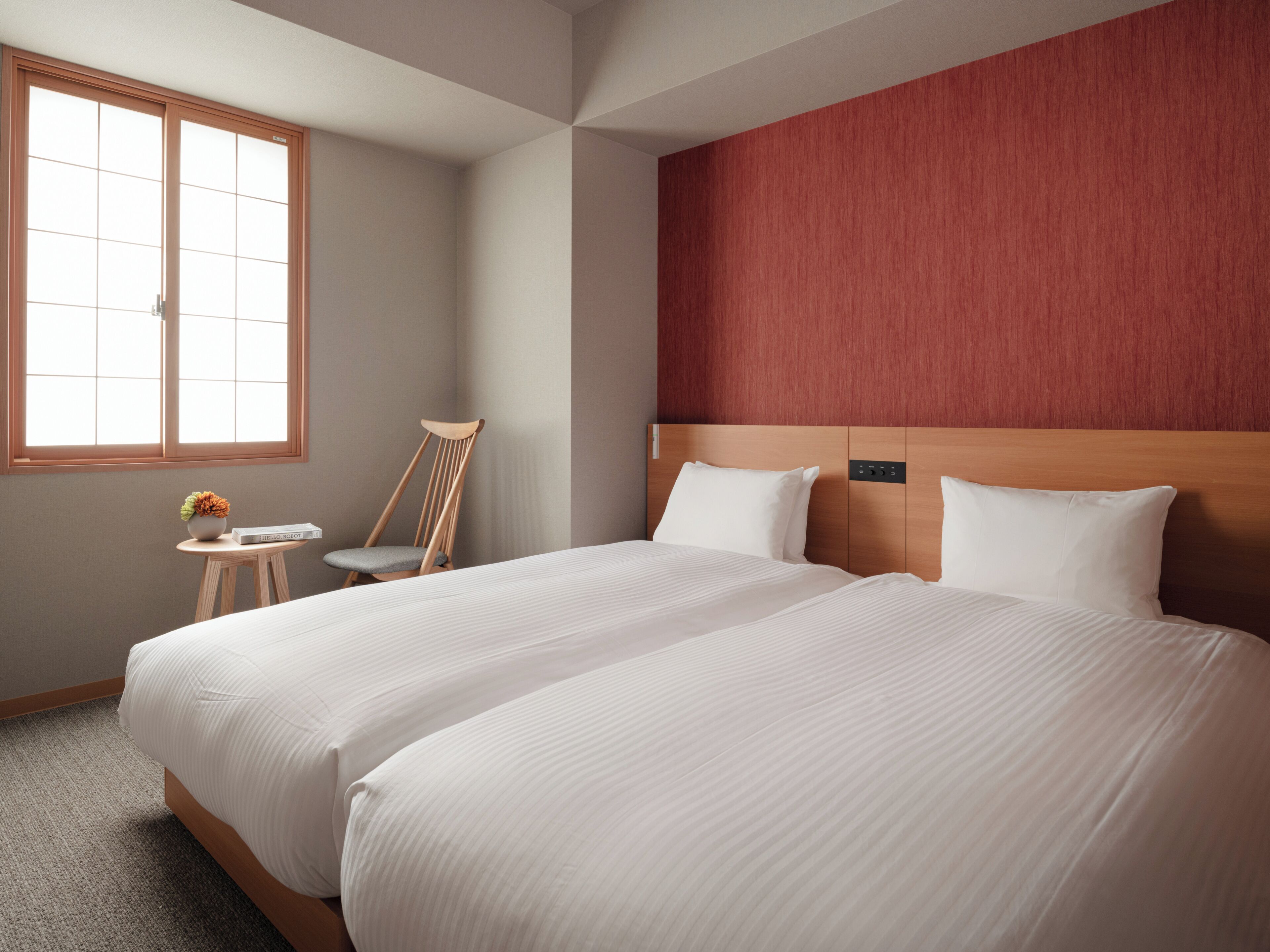 Une chambre d’hôtel avec deux lits, une tête de lit en bois, une petite table avec un vase et une fenêtre avec des stores.