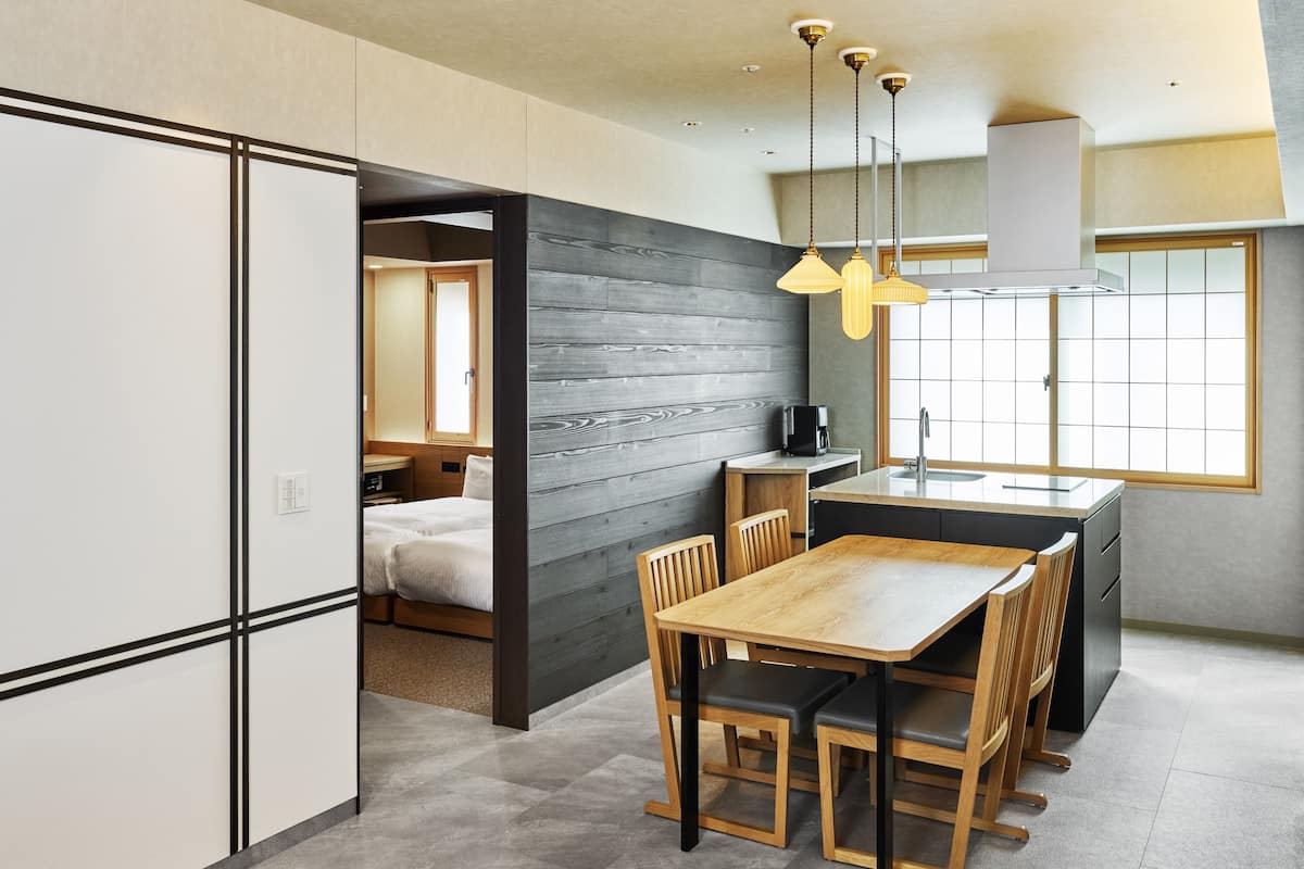 A modern kitchen with a dining area, featuring a wooden table and chairs, a built-in stove, and a large window providing natural light.
