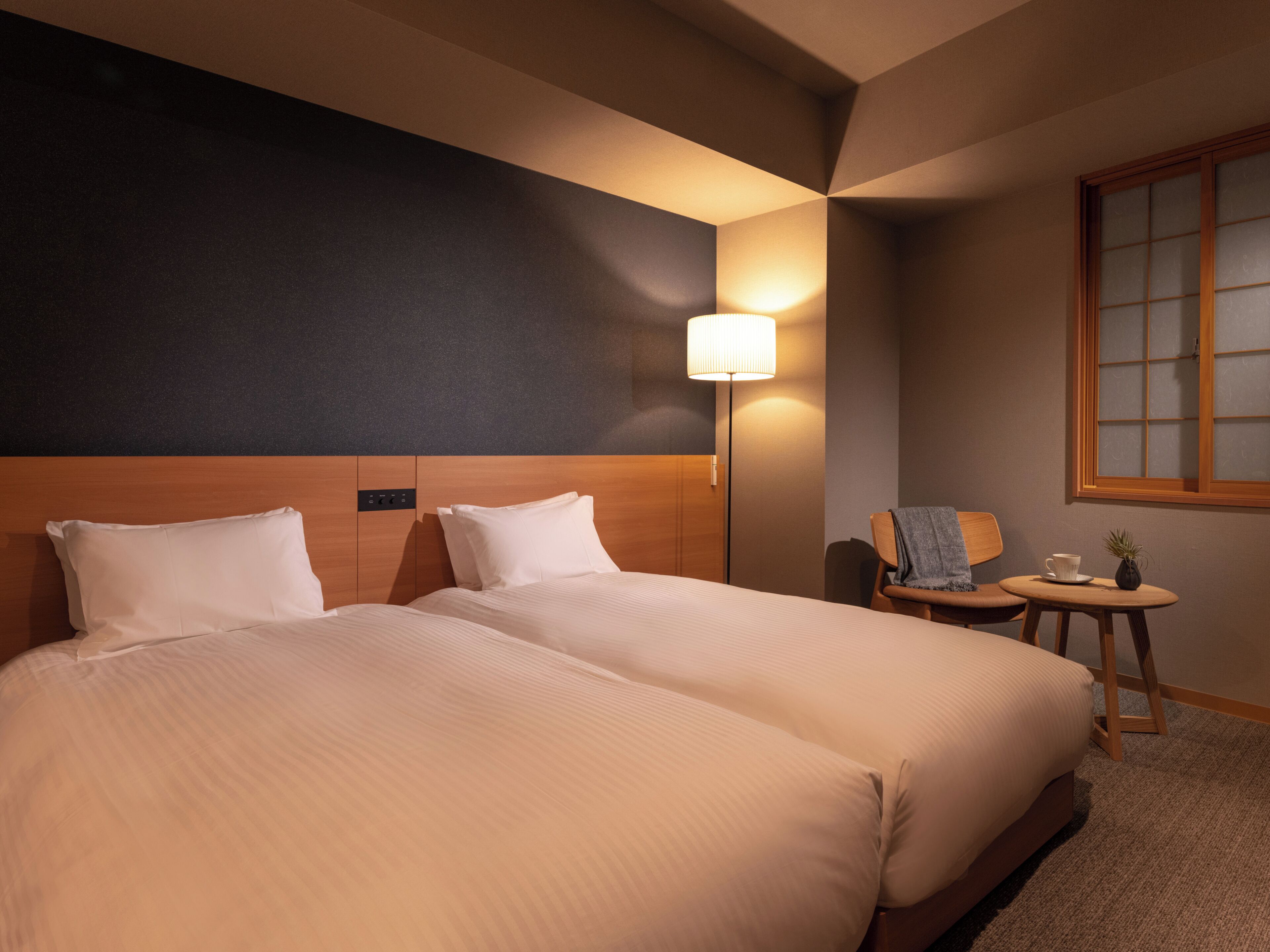 Une chambre d’hôtel avec deux lits, une table de chevet, une lampe, une chaise et une petite table sur laquelle se trouvent une tasse et une plante.