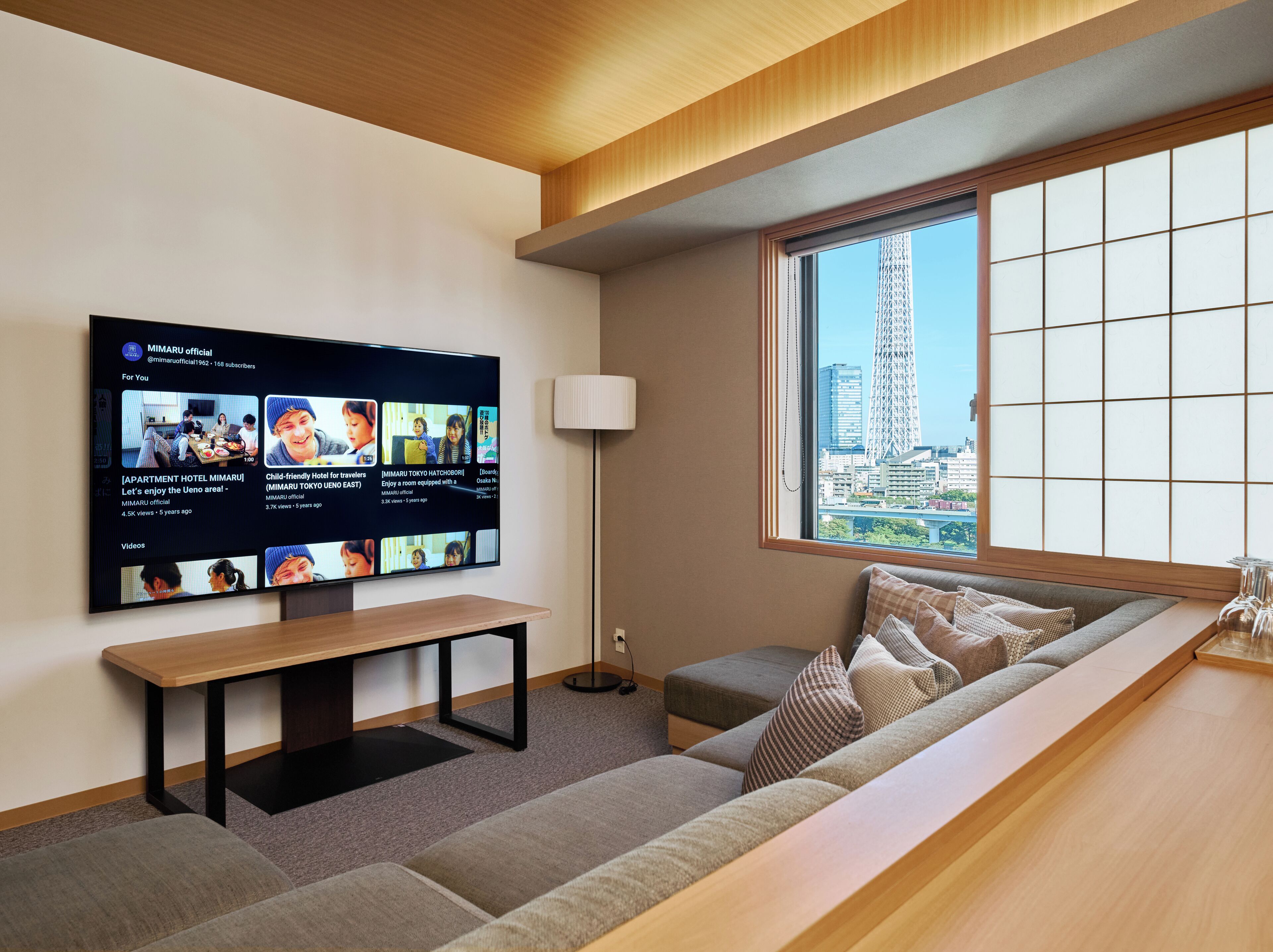 A modern living room with a large flat-screen TV mounted on the wall, a wooden bench, a sofa with cushions, a floor lamp, and a large window with a city view.