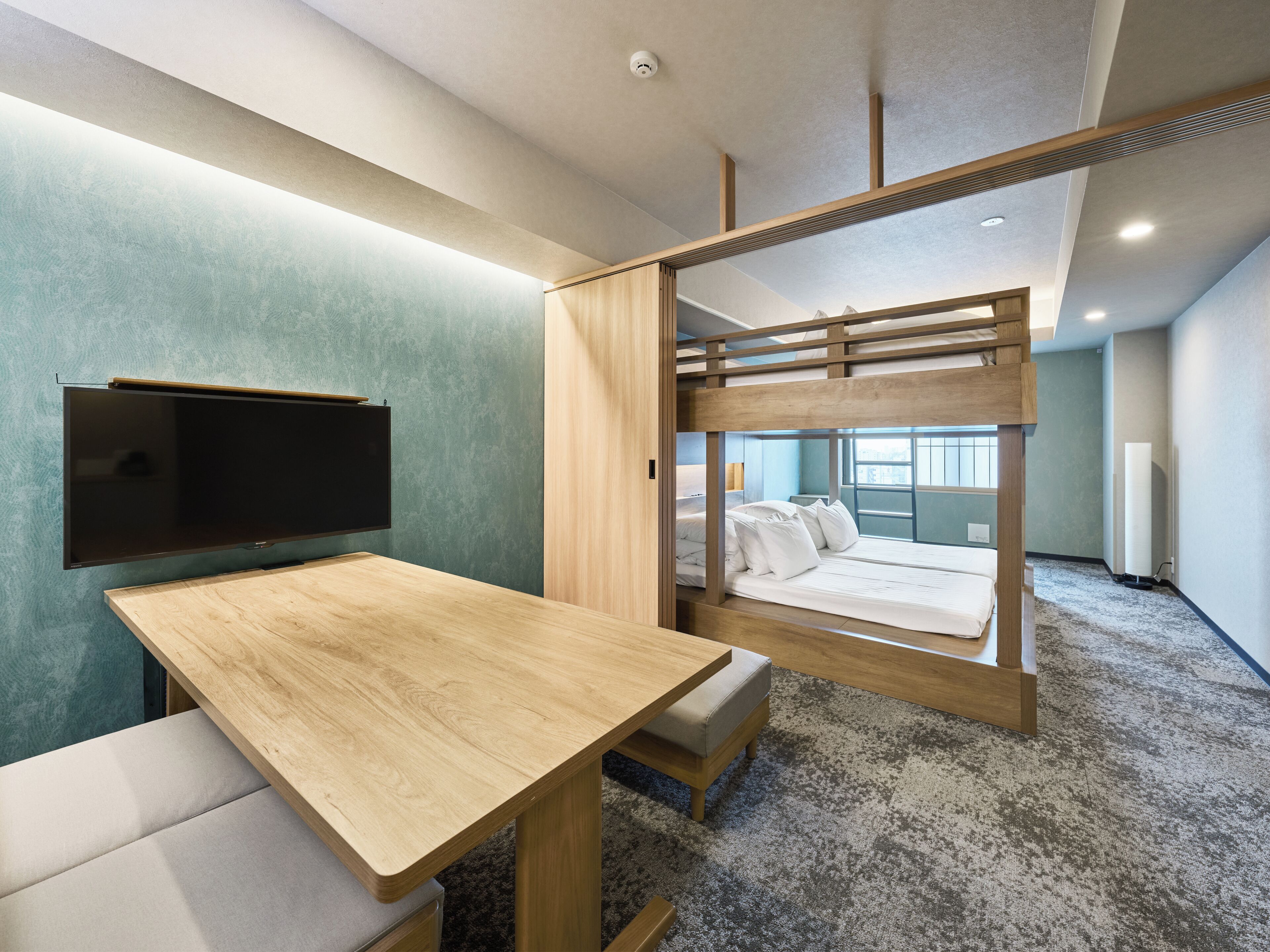 A bunk bed with a TV mounted on the wall, a wooden desk, and a cushioned bench.