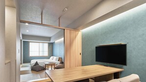 A modern hotel room with a wooden dining table, a sofa, a TV mounted on the wall, and a sliding door leading to a balcony.