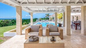 A covered outdoor seating area with a pool, wicker furniture, and a view of the ocean.
