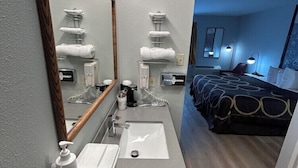 A modern bathroom with a large mirror, a sink, and a toilet. There is a bed with a blue wall and a nightstand in the adjacent room. - Super 8 By Wyndham Sterling CO (Sterling)