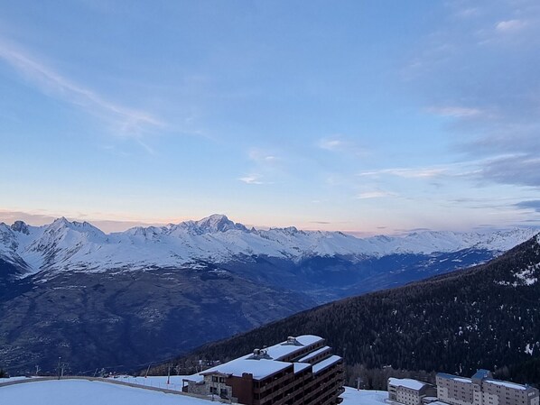 A snow-covered mountain range with a clear sky. There are buildings with multiple floors and a flat roof, likely part of a hotel or resort.