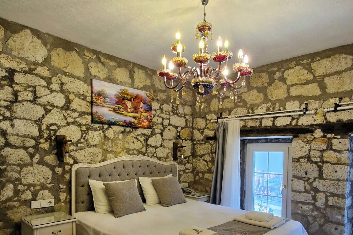Ein Schlafzimmer mit Steinwand, einer Chandelier, einem Bett mit Kopfteil, einem Nachttisch und einem Fenster mit Vorhängen.