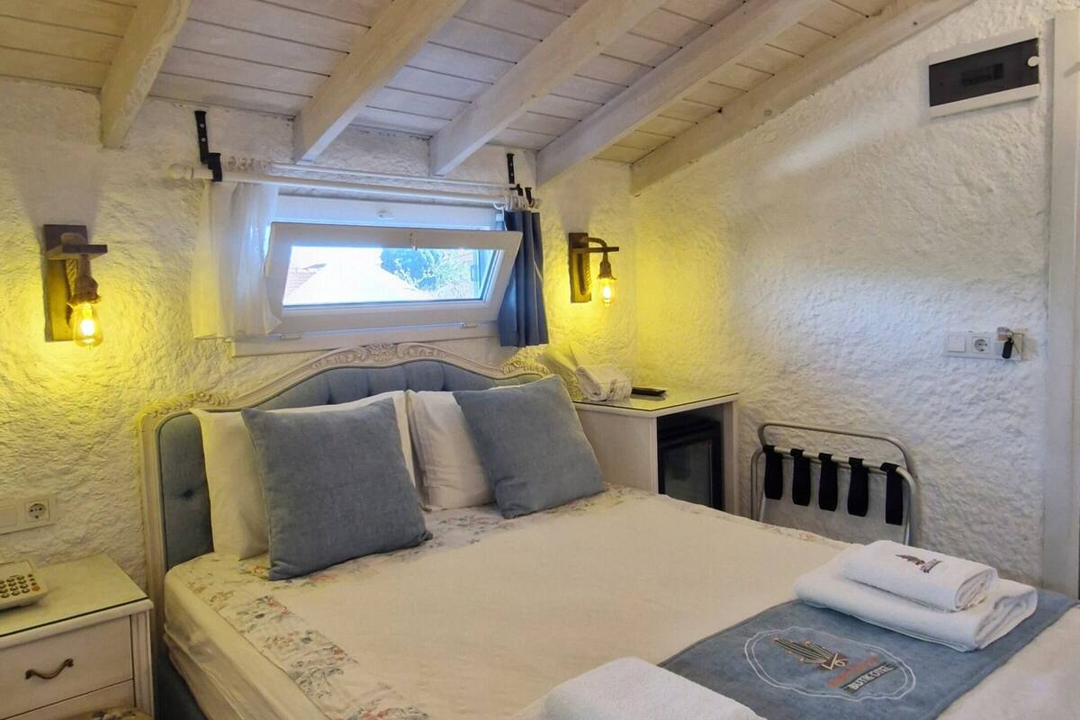 Ein Schlafzimmer mit Holzbalkendecke, einem Bett mit Blumenmuster-Bettwäsche, einem Nachttisch, einem Fenster mit Vorhängen und einer wandmontierten Lampe.