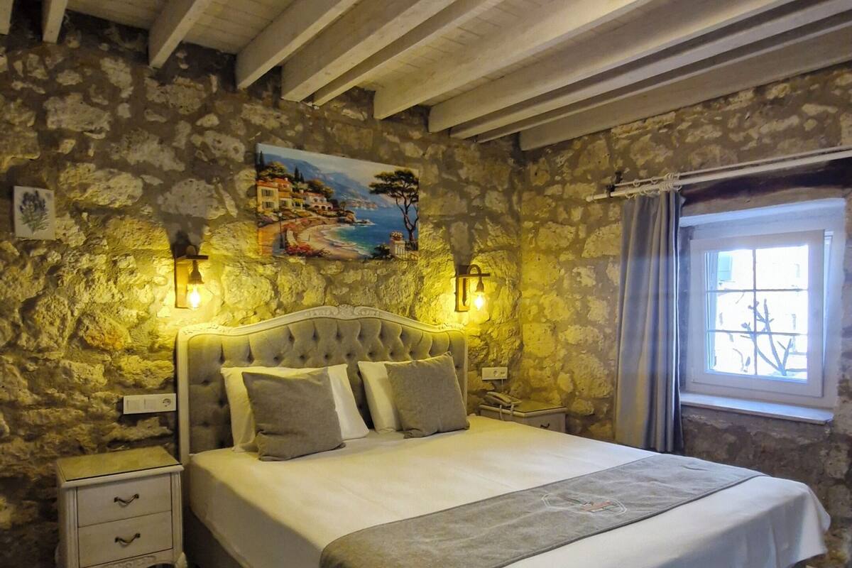 Ein Schlafzimmer mit Steinwand, einem Bett mit grauem Kopfteil, einem Nachttisch, einem Fenster mit Vorhängen und einem Bild an der Wand.