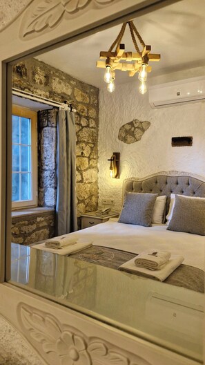 Une chambre avec un mur en pierre, un lit doté d’une tête de lit capitonnée, une table de chevet avec une lampe et une fenêtre avec des rideaux.