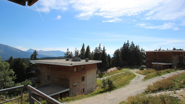 A rustic building with a brick exterior and a flat roof, surrounded by a gravel path and dense forest.