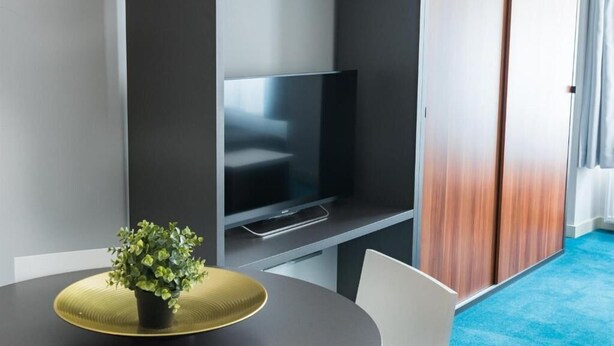 A modern hotel room with a flat-screen TV mounted on a wall, a small potted plant on a round table, and a blue carpet. - Cilindar Apartments (Zagreb)