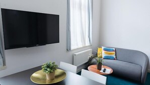 A modern living room with a flat-screen TV mounted on the wall, a grey sofa with colorful cushions, a small round table with a plant, and a dining table with a gold-rimmed plate. - Cilindar Apartments (Zagreb)