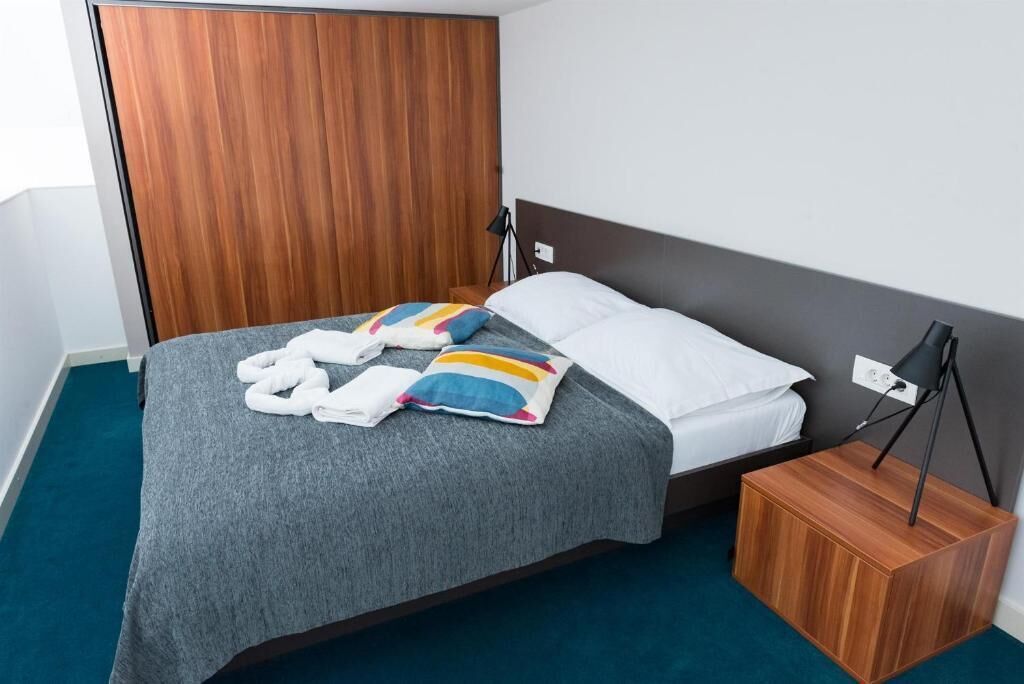 A neatly made bed with a grey blanket, a colorful pillow, and a pair of white towels. There is a wooden nightstand with a lamp beside the bed.