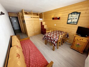 Quarto compacto com beliche, mesa de jantar com cadeiras, televisão em móvel de madeira e quadro pendurado na parede.