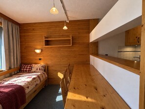 A compact room with a bed, a wooden bench, a small kitchen area, and a wall-mounted shelf.