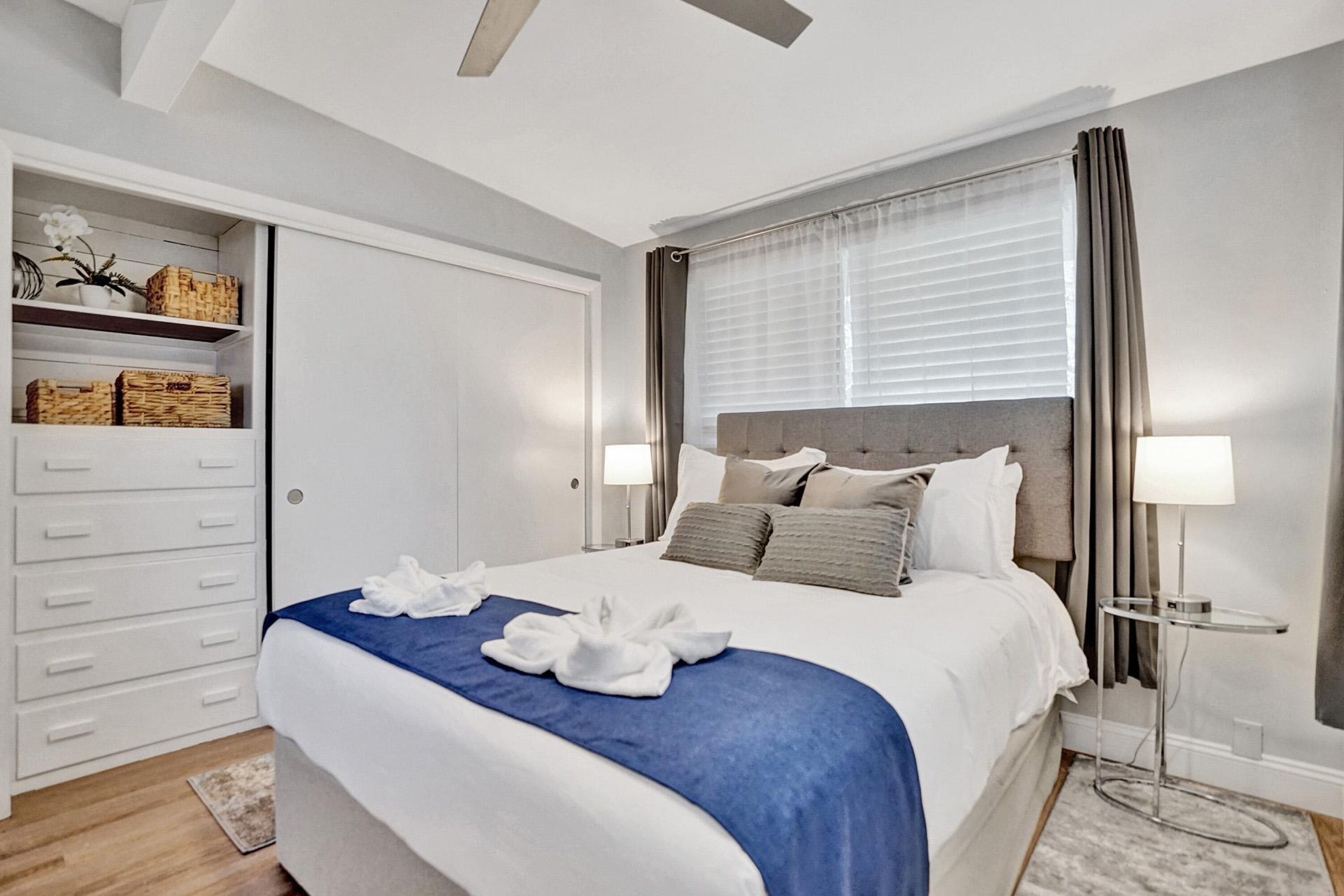 Quarto de hotel moderno com cama bem arrumada, mesa de cabeceira com luminária, guarda-roupa e janela com cortinas.