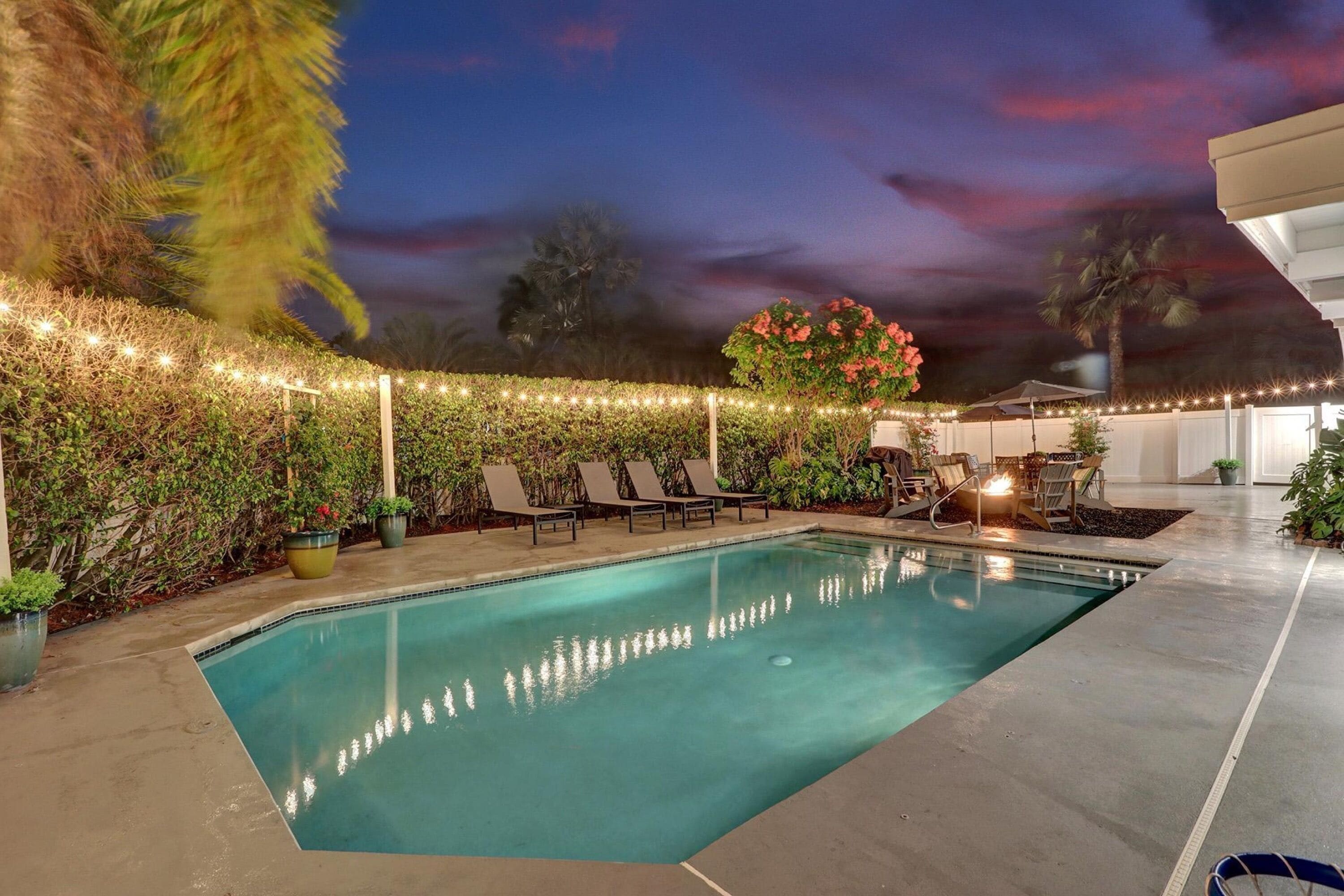 An outdoor swimming pool with lounge chairs, string lights, and a well-maintained hedge.
