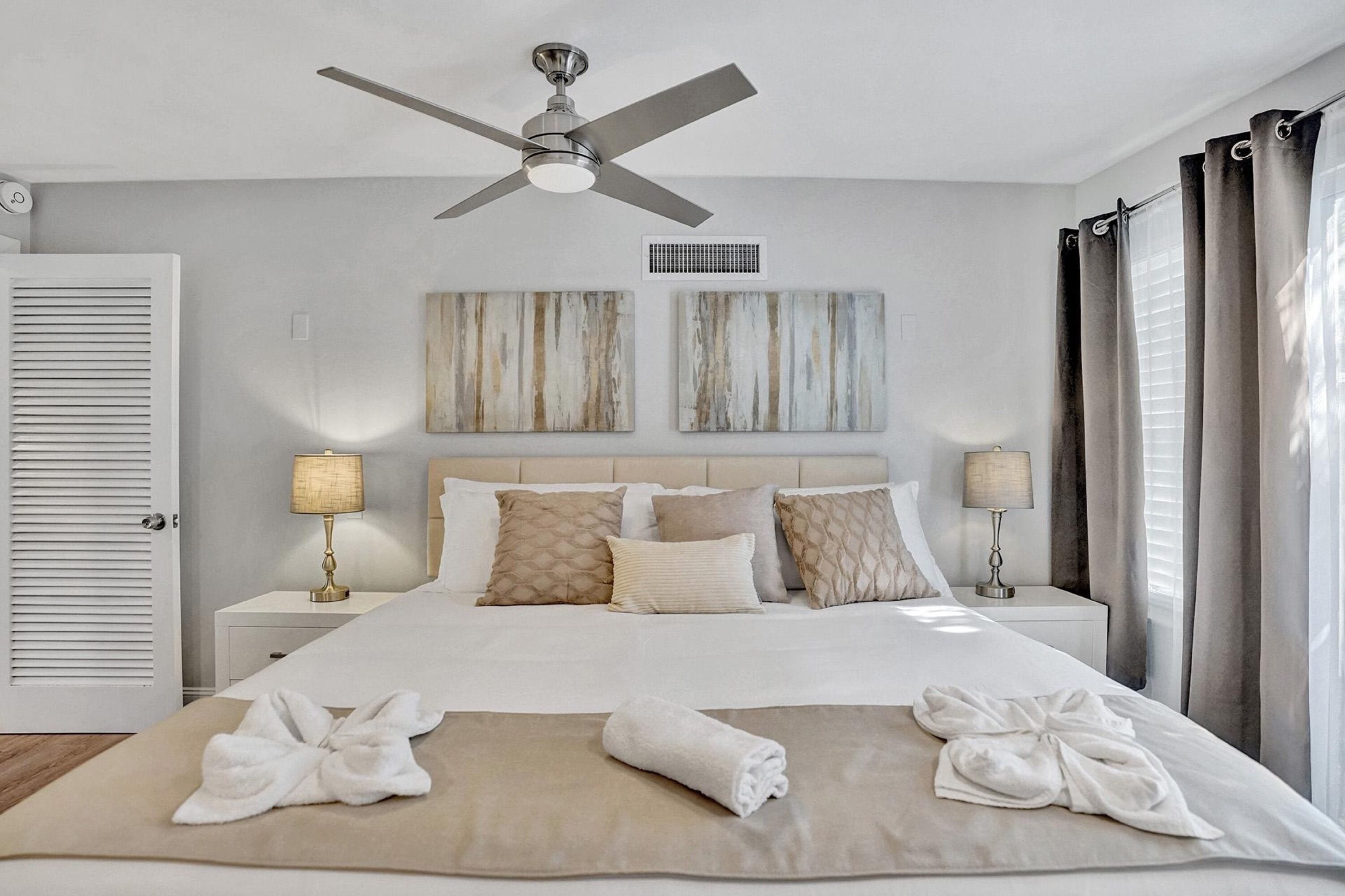 Un lit bien fait avec deux oreillers, une table de chevet avec une lampe, un ventilateur de plafond et des œuvres d’art accrochées au mur.