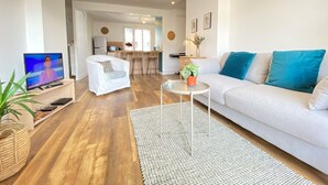 A modern living room with a white sofa, a coffee table, and a TV. There is a kitchen area in the background with a fridge, microwave, and bar stools. - Renovated T3 Apt with Balcony & Parking in Annecy Center (Annecy)