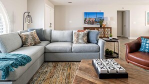 A living room with a grey sectional sofa, a coffee table with a wooden base, a patterned rug, and a wall-mounted TV. - Art Deco Oasis: Vintage Charm, Pool, Firepit, BBQ (Scottsdale)