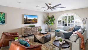 A living room with a ceiling fan, a large window, a sofa, a coffee table, and a TV. - Art Deco Oasis: Vintage Charm, Pool, Firepit, BBQ (Scottsdale)