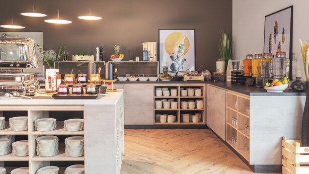 A modern kitchen with a central island, wooden cabinets, and a variety of food items and utensils. - Candlewood Suites Augsburg by IHG (Gersthofen)