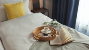 A neatly made bed with a tray holding a cup and a book, a lamp, and a plant on a bedside table. - Candlewood Suites Augsburg by IHG (Gersthofen)