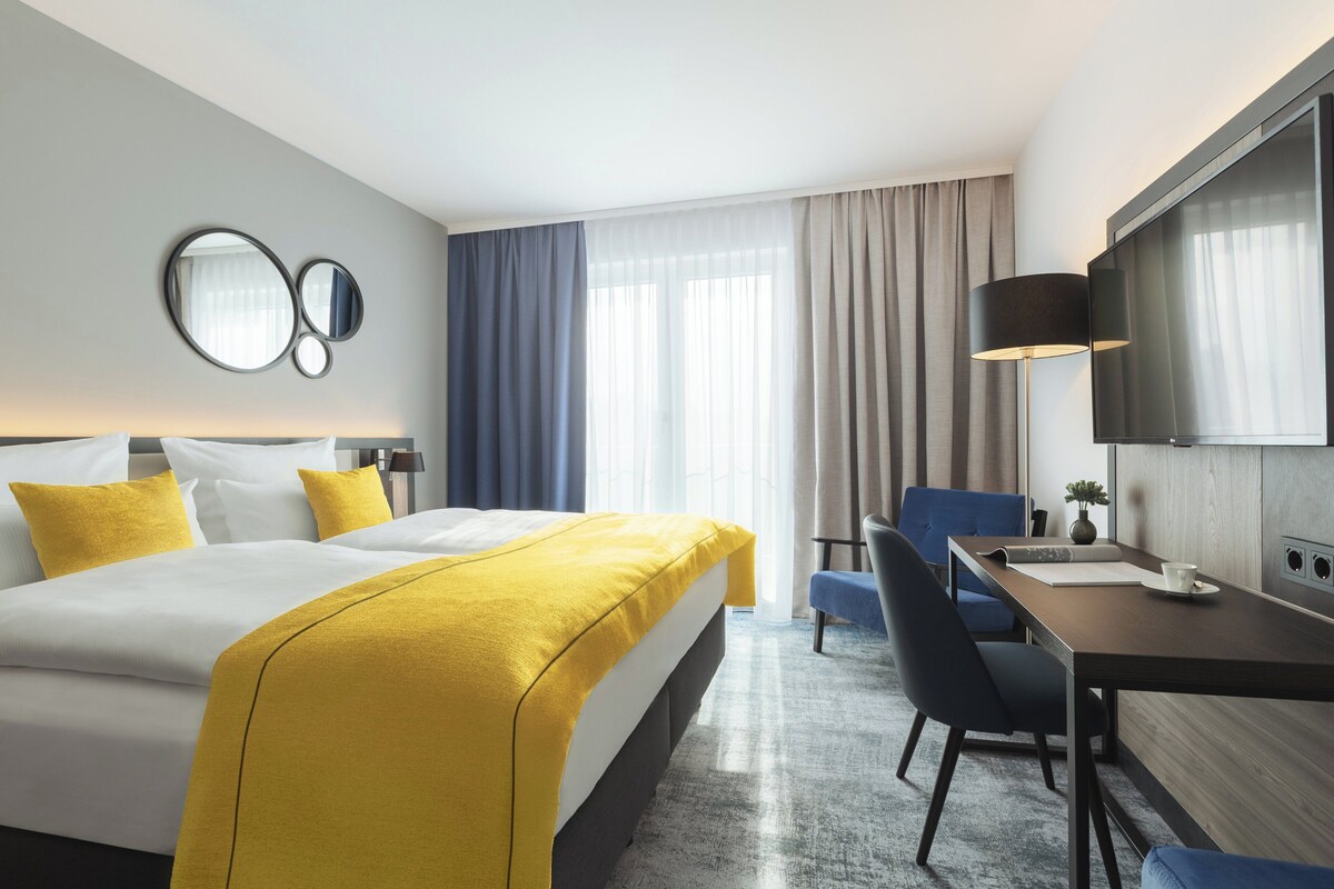 A hotel room with a bed, a desk, a chair, a TV, and a large window with curtains.