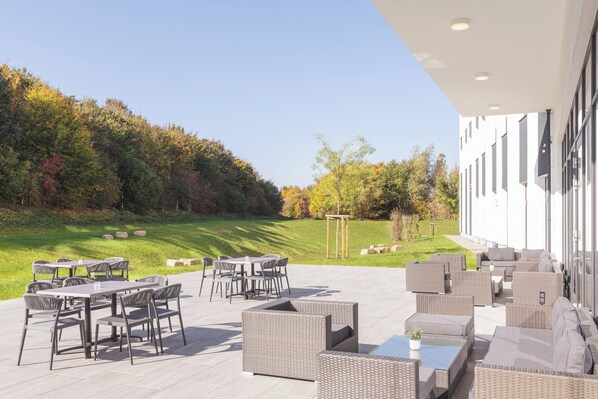 An outdoor patio with wicker furniture, a round table, and a view of a grassy area with trees. - Candlewood Suites Augsburg by IHG (Gersthofen)