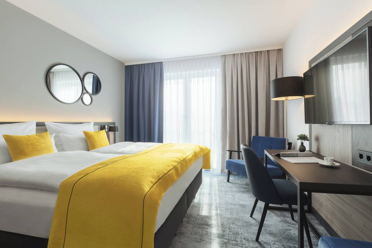 Ein Hotelzimmer mit Bett, Schreibtisch, Stuhl, Fernseher und einem großen Fenster mit Vorhängen.