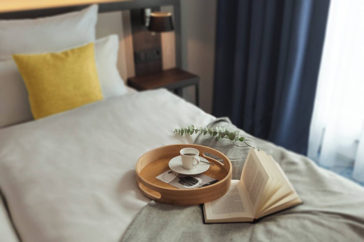Ein ordentlich bezogenes Bett mit einem Tablett, das eine Tasse und ein Buch hält, einer Nachttischlampe und einem Fenster mit Vorhängen.