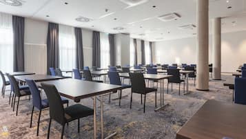 A conference room with multiple tables and chairs, large windows with curtains, and a carpeted floor.