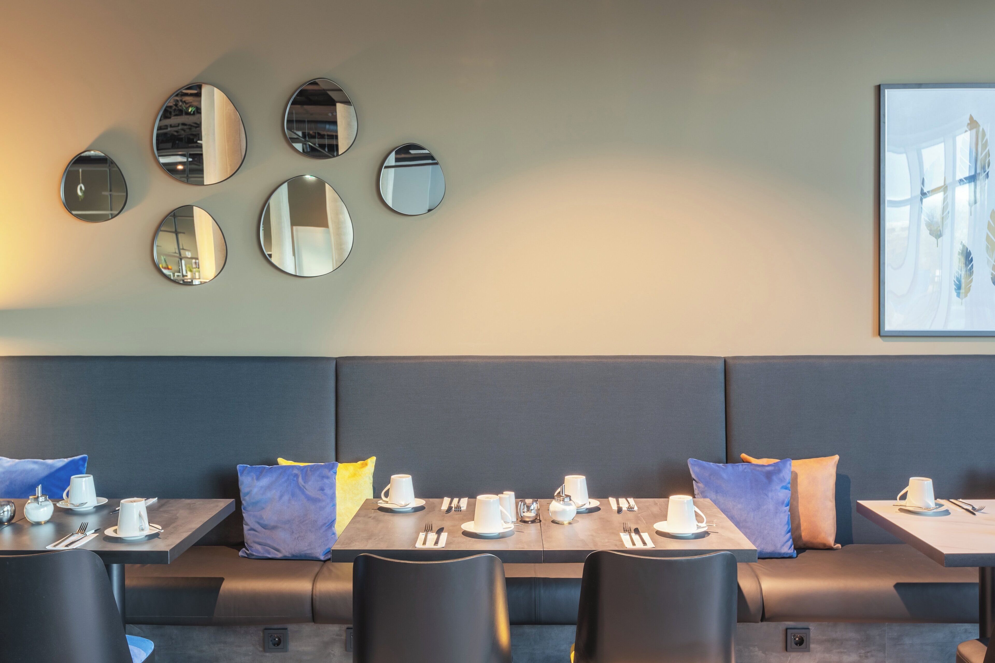 Un comedor moderno con espejos redondos en la pared, un banco acolchado azul, y mesas dispuestas con tazas y cubiertos blancos.