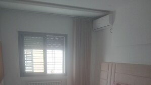 Quarto com cama grande, janela com persianas, radiador e ar condicionado fixado na parede.