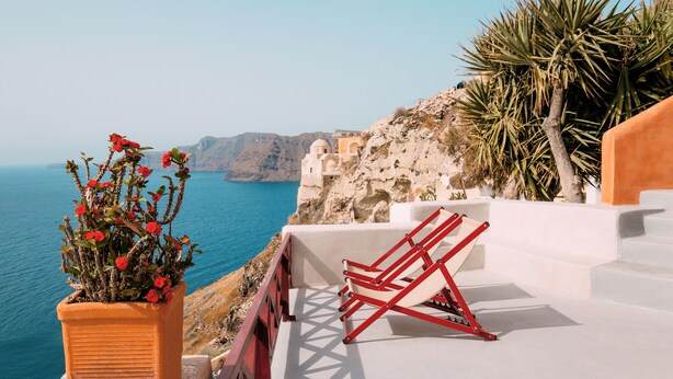 A seaside terrace with a view of the sea, a potted plant with red flowers, and red lounge chairs. - Terracotta Cave House (1) (Santorini)