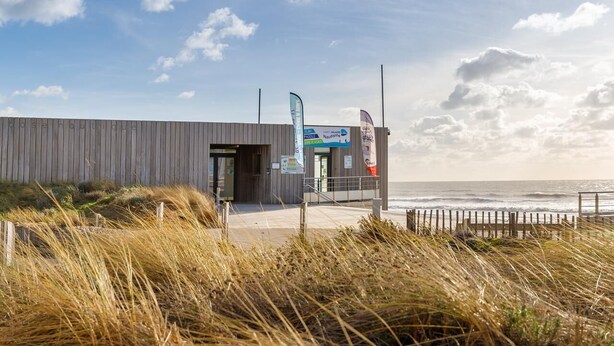 Ein modernes Gebäude mit Holzfassade in Strandnähe, mit einer Schautafel und Flaggen.
