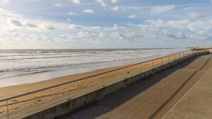 Eine Uferpromenade mit Betonweg entlang des Strandes, der zu einem Steg mit Gebäuden in der Ferne führt.