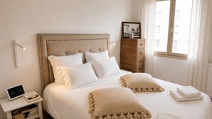 A neatly made bed with a headboard, a bedside table with a clock, a wooden dresser, and a window with curtains.
