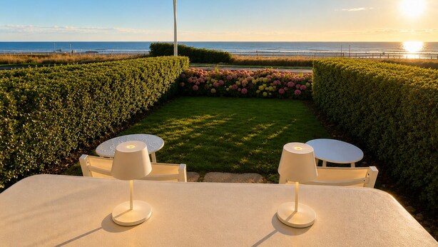 A seaside patio with two round tables, a lamp, and a hedge.