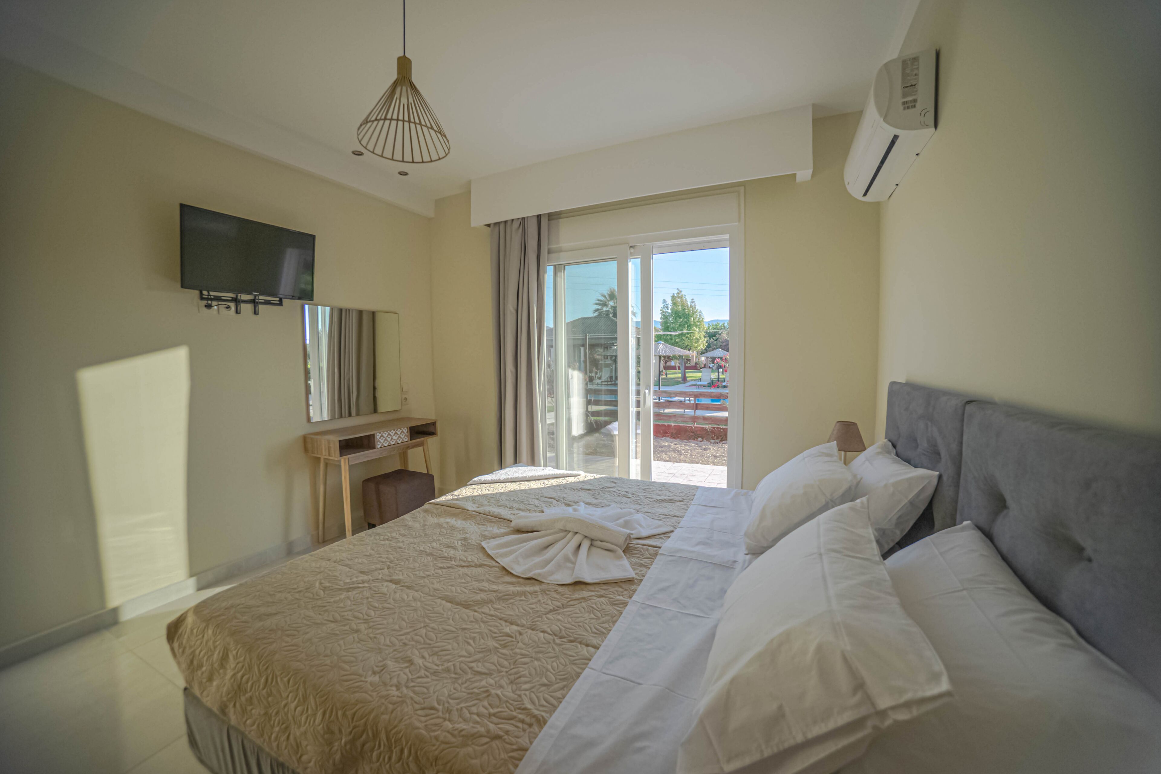 A hotel room with a bed, a flat-screen TV mounted on the wall, a desk with a lamp, and a sliding glass door leading to an outdoor area.