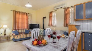 A cozy living room with a sofa, a dining table with fruit and drinks, a TV, and a kitchen area with cabinets.