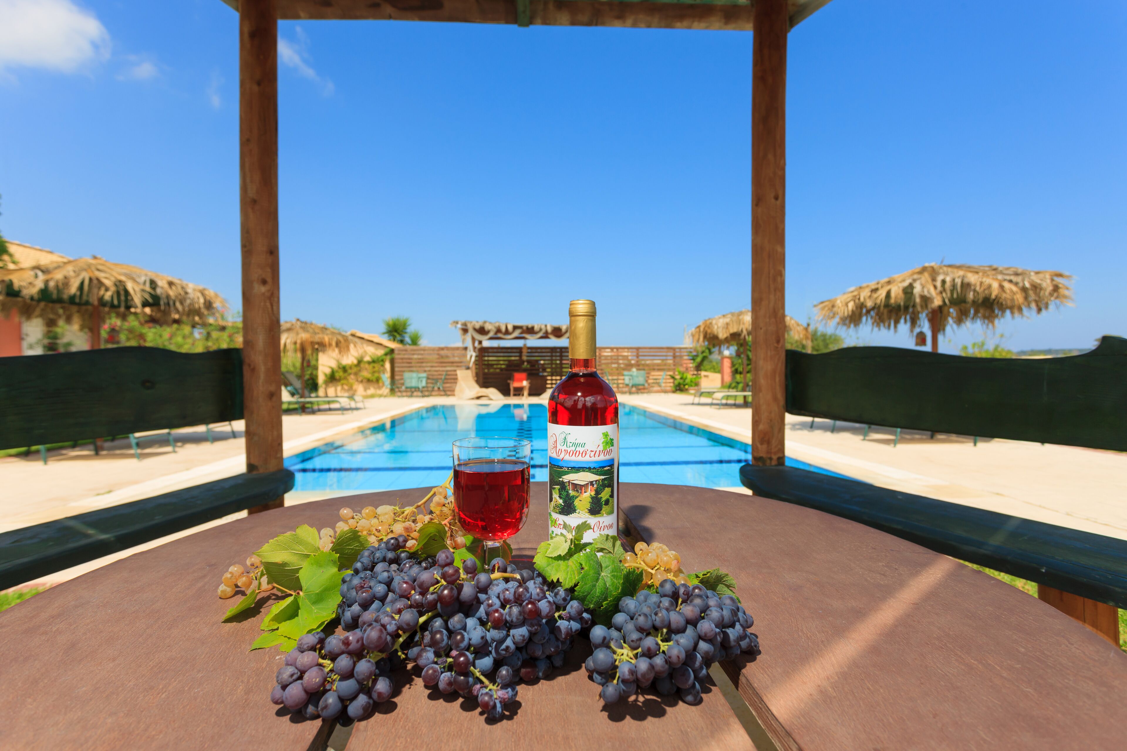 Une table en bois sur laquelle se trouvent une bouteille de vin rouge, un verre de vin rouge et une grappe de raisins, avec une piscine en arrière-plan.