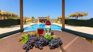 A table with a bottle of red wine, a glass of red wine, and a bunch of grapes on a wooden table with a pool in the background.