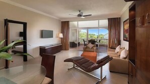 A modern hotel room with a glass dining table, a leather sofa, a reclining chair, a TV, and a balcony with a view.