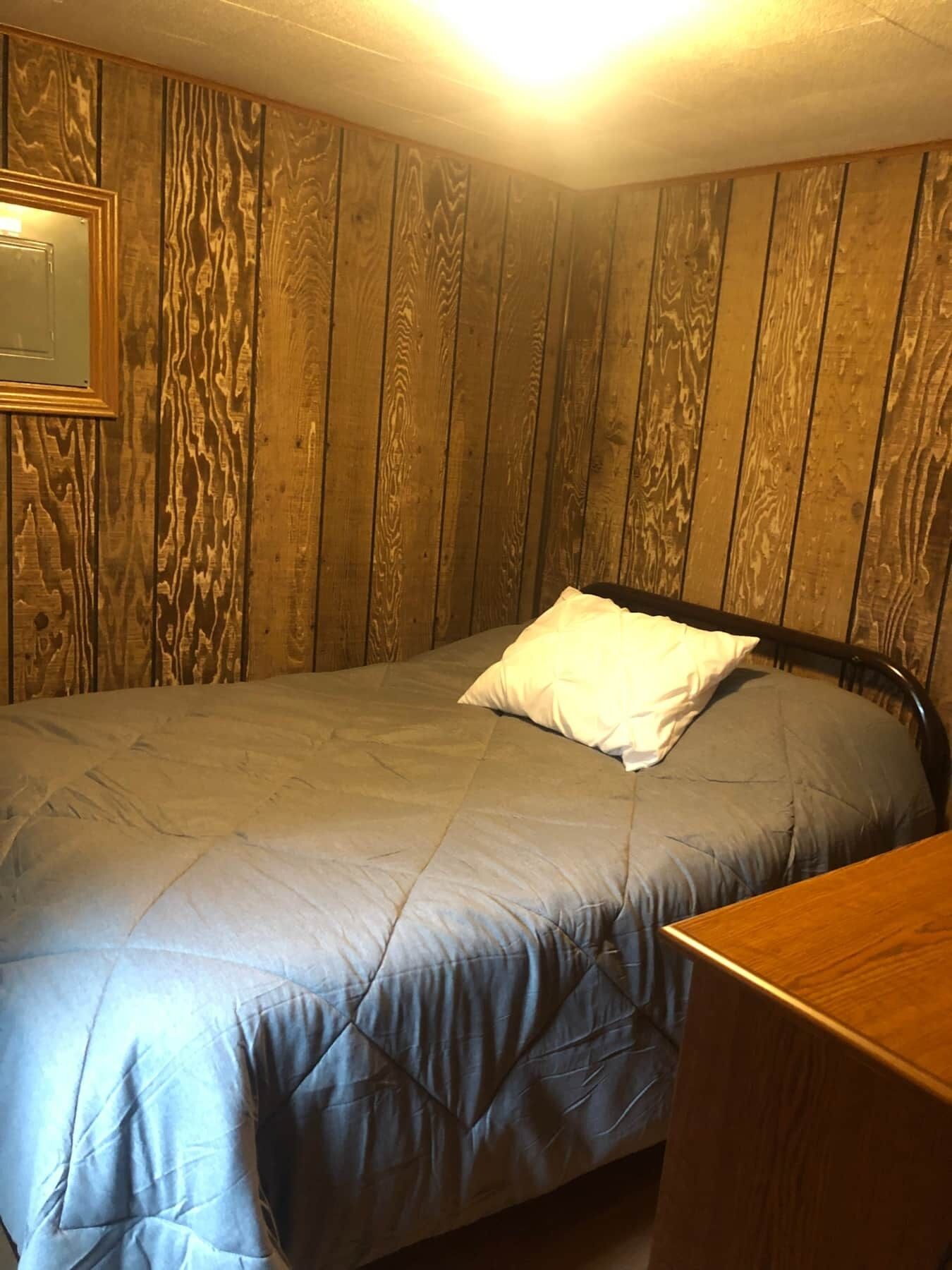 Un dormitorio con paneles de madera, una cama con edredón gris y almohada blanca, y una mesita de noche de madera.
