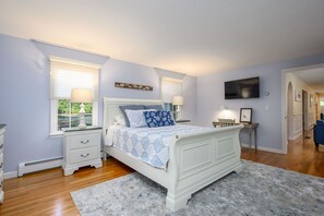 Une chambre avec un lit blanc, deux tables de chevet avec des lampes, un téléviseur à écran plat fixé au mur et une fenêtre avec des stores.