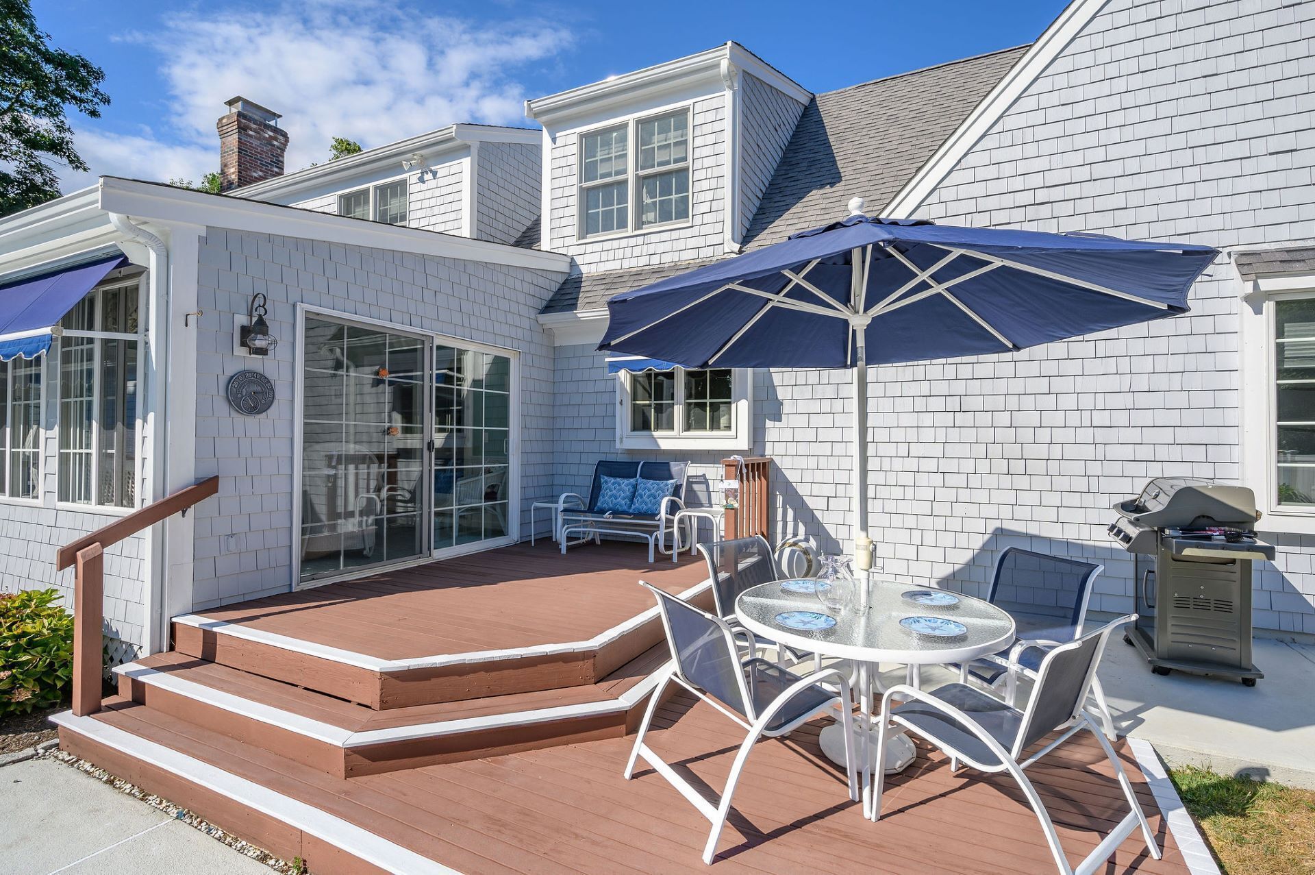 Ein Haus mit einer Terrasse, inklusive Tisch und Stühlen, einem Grill und einem überdachten Bereich mit einem blauen Sonnenschirm.